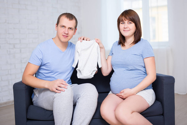 conceito de família jovem - belo casal grávida sentado no sofá na sala de estar e segurando bodysuit bebê
 - Foto, Imagem