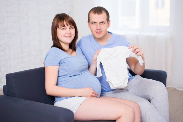 conceito de família - jovem belo casal grávida sentado no sofá na sala de estar e segurando bodysuit bebê
 - Foto, Imagem