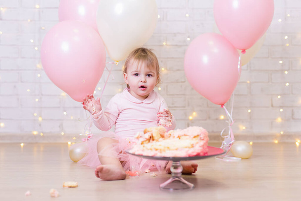primera celebración de cumpleaños - niña divertida con pastel roto sobre fondo de pared de ladrillo con luces y globos de colores
 - Foto, imagen
