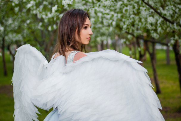 Portret młodej, pięknej kobiety w białym gorsecie i z dużymi białymi anielskimi skrzydłami za plecami na tle kwitnących jabłoni w parku. Anioł w rajskim ogrodzie. - Zdjęcie, obraz