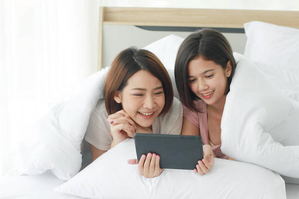 アジアの若い姉妹の素敵なカップルは、白いベッドの上に座って、ベッドルームでノートパソコンやスマートフォンと一緒にソーシャルオンラインで笑顔。同性愛者とアジア系レズビアンの関係。テクノロジーコンセプト - 写真・画像