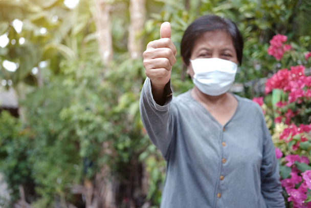 Asian old woman wearing a white cloth mask for prevent the Covid-19 or Corona virus in Thailand and Thumbs up for protection of Air Pollution. Health of elderly concept. Closed up focus on hand. - Photo, Image