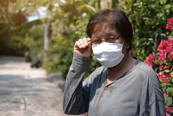 Asian old woman wearing glasses and white cloth mask for prevent the Covid-19 or Corona virus in Thailand and protect for Air Pollution Value Pm 2.5. Health and medicine of elderly in garden concept - Photo, Image