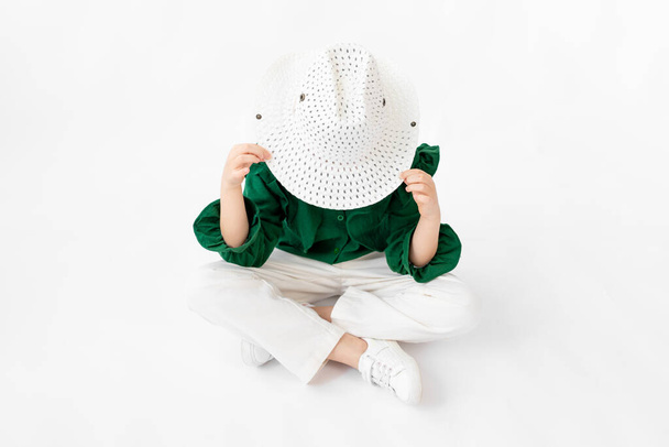 a girl sits on a white background and hides behind a hat, space for text - Foto, afbeelding