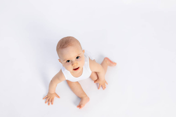 Leuke peuter jongen 8 maanden oud in wit bodysuit zit op witte achtergrond, kijken naar de bovenkant, ruimte voor tekst - Foto, afbeelding