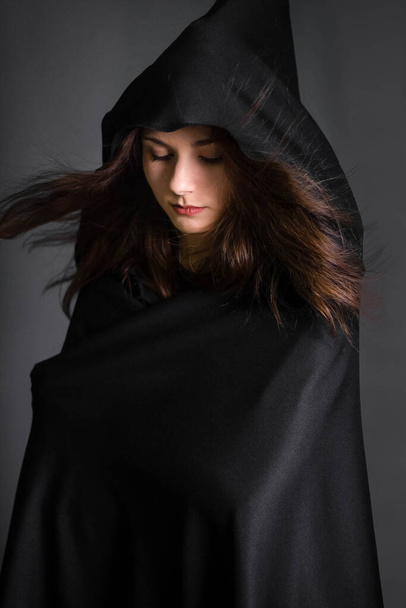 Young, beautiful woman in a black cloak with a hood in the studio on a gray background. Long hair fluttering on the wind. A model with clean skin. Suit of a fortuneteller, witches for Halloween. - Fotografie, Obrázek