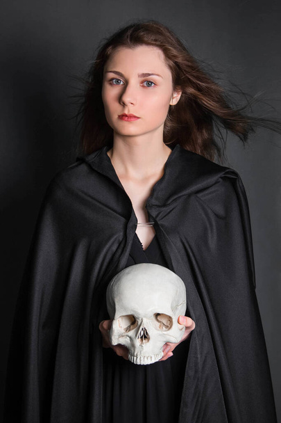 Mooie, jonge vrouw met lang haar in een zwarte mantel met een schedel in haar handen. Studio foto op een grijze achtergrond. Hekserij, necromantie, waarzeggerij, Halloween. - Foto, afbeelding