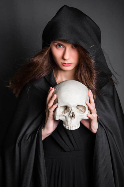 A beautiful, young woman with long hair in a black cloak with a hood, with a skull in her hands. Studio photo on a gray background. Witchcraft, necromancy, divination, Halloween. - Valokuva, kuva