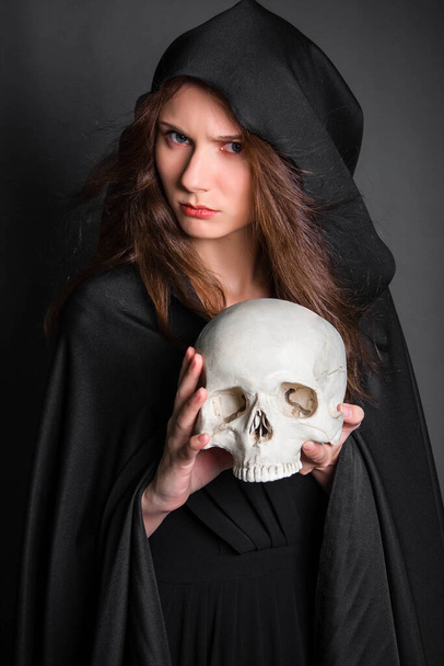 A beautiful, young woman with long hair in a black cloak with a hood, with a skull in her hands. Studio photo on a gray background. Witchcraft, necromancy, divination, Halloween. - Φωτογραφία, εικόνα