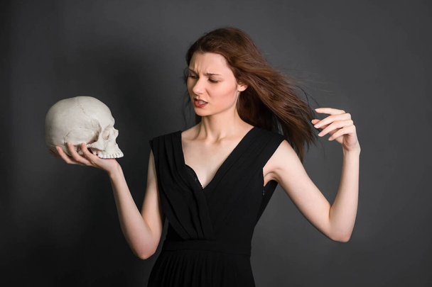 A beautiful, young woman with long hair in a black dress with a skull in her hands. Studio photo on a gray background. Witchcraft, necromancy, fortune telling, Halloween. - Фото, изображение