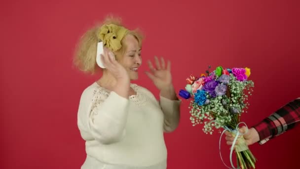 Uśmiechnięta starsza kobieta słuchająca muzyki i przyjmująca kwiaty z uśmiechem - Materiał filmowy, wideo