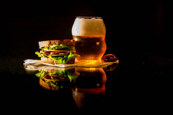 A glass of beer and a sandwich with cheese, sausage and salad on a napkin are on a glass table with reflection on a black background - Foto, afbeelding
