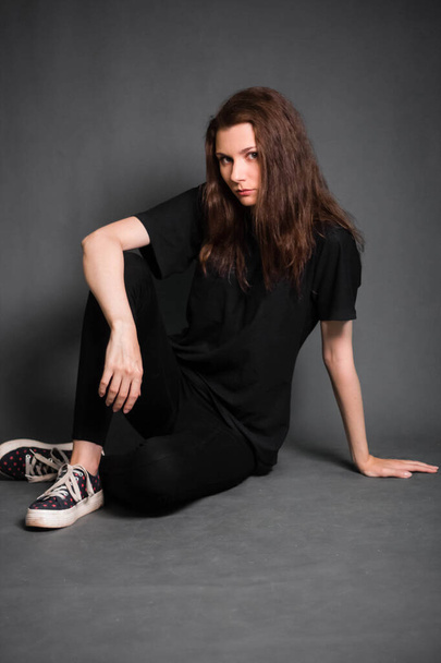 Beautiful, young woman with long hair, in black clothes is sitting on the floor. Studio photo on a gray background. A model with clean skin. - Zdjęcie, obraz