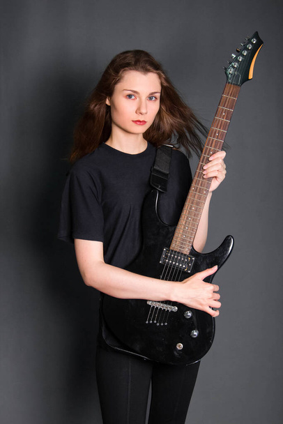 Portrait of a beautiful, young rock woman in black clothes with an electric guitar in her hands. Studio photo on a gray background. Model with clean skin. - Foto, Bild