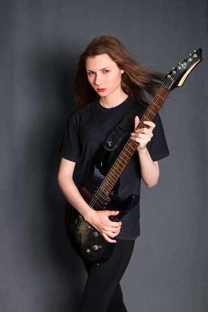 Πορτρέτο μιας όμορφης, νεαρής ροκ γυναίκας με μαύρα ρούχα και ηλεκτρική κιθάρα στα χέρια της. Φωτογραφία στούντιο σε γκρι φόντο. Μοντέλο με καθαρό δέρμα. - Φωτογραφία, εικόνα