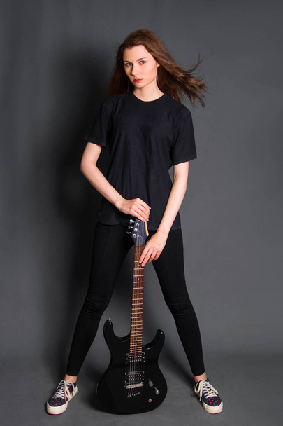 Attractive, young rock woman in black clothes with an electric guitar in her hands stands on a gray background. Model with clean skin. - 写真・画像