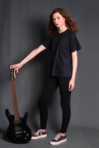 Attractive, young rock woman in black clothes with an electric guitar in her hands stands on a gray background. Model with clean skin. - Photo, image
