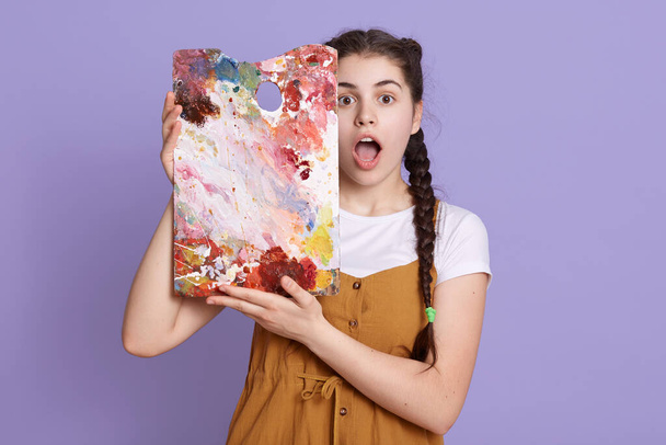Indoor shot of creative young woman painting in her studio. Female artist in her workshop holding colors palette near her face, stands with widely open mouth, expresses shock, poses against lilac wall - Fotografie, Obrázek