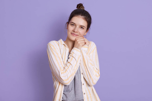 Charming dark haired girl with bun, standing against lilac wall and keeping hands together near face, wearing casual attire, cute lady looks happy with smile. - Foto, Imagen