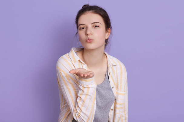 Charming brunette girl wearing casual clothing showing air kiss, keeps lips rounded, standing isolated over lilac background, female looking at camera. - Fotó, kép