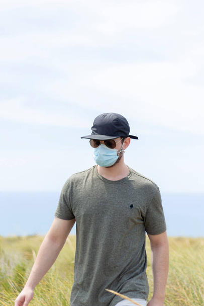 Un hombre caminando por la vegetación con una máscara facial, una gorra y un par de anteojos en un día soleado
 - Foto, imagen