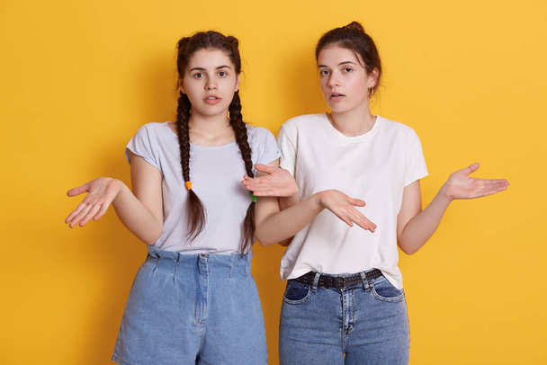 Two young teenage girls in white t shirts posing with spreading hands, showing helpless gesture, have astonished facial expressions, keep mouths opened. - Foto, Bild