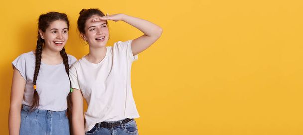 Happy smiling girl keeping palm near forehead and looking far away, standing against yellow wall, girl with dark hair wearing white t shirt. Copy space for advertisement. - Foto, Imagen