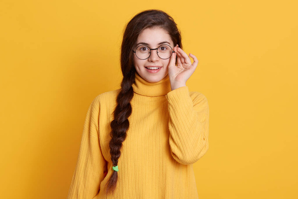 Happy smiling excited girl with pigtail wearing yellow shirt, touching frame of her glasses, being in good mood, expressing positive emotions,looks satisfied. - Zdjęcie, obraz