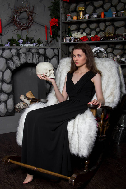 Молодая красивая девушка в черном платье с черепом в руках, сидит в кресле-качалке возле камина. Модель с чистой кожей. Привлекательная ведьма. Хэллоуин
. - Фото, изображение