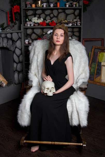 A young, beautiful woman in a black dress with a golden crown on her head and a skull in her hands is sitting in a rocking chair near the fireplace. A model with clean skin. Attractive witch, Halloween. - 写真・画像