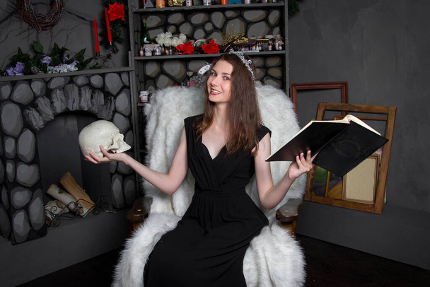 Een jonge, mooie vrouw in een zwarte jurk met een zilveren diadeem op haar hoofd, met een schedel en een boek in haar handen, zit in een schommelstoel bij de open haard. Hij leest een spreuk, tovert. Halloween. - Foto, afbeelding