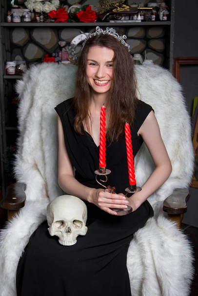 A young, beautiful woman in a black dress with a silver diadem on her head, with a skull and candles in her hands, sits in a rocking chair near the fireplace. A model with clean skin. witch. Halloween. - Foto, afbeelding