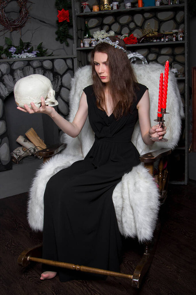 A young, beautiful woman in a black dress with a silver diadem on her head, with a skull and candles in her hands, sits in a rocking chair near the fireplace. A model with clean skin. witch. Halloween. - Foto, afbeelding