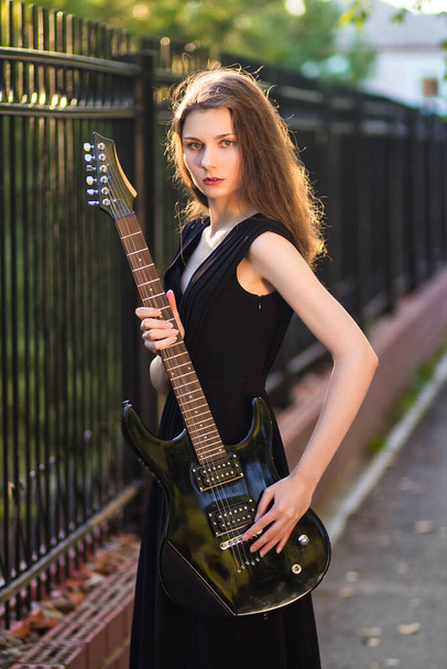 Beautiful, young woman in a black dress and with a guitar in her hands, against a background of steel fence and trees. - Photo, Image