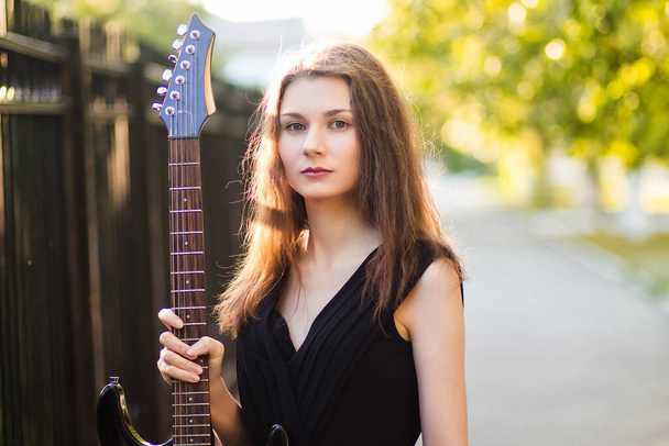 Beautiful, young woman in a black dress and with a guitar in her hands, against a background of steel fence and trees. - Fotoğraf, Görsel