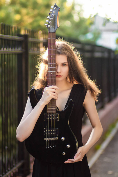 Beautiful, young woman in a black dress and with a guitar in her hands, against a background of steel fence and trees. - Foto, afbeelding