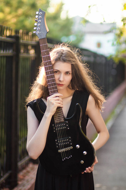 Bella, giovane donna in abito nero e con una chitarra tra le mani, su uno sfondo di recinzione in acciaio e alberi. - Foto, immagini