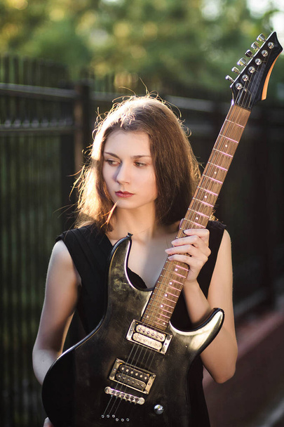 Beautiful, young woman in a black dress and with a guitar in her hands, against a background of steel fence and trees. - Фото, изображение