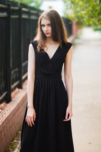 young, beautiful woman in black dress, outdoor fashionable photo on a background of steel fence, summer evening. Moddel with clean skin. - 写真・画像