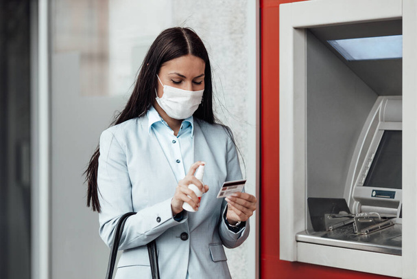 Elegant woman with protective mask standing on city street and using alcohol spray to disinfect her hands after use of ATM machine. Corona or Covid-19 virus pandemic prevention and healthcare concept. - Фото, изображение