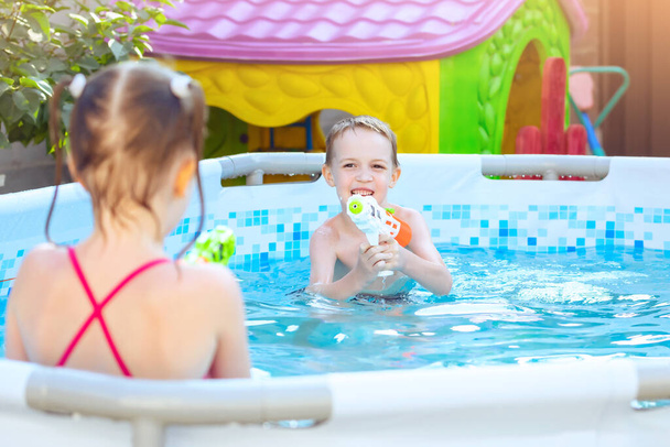 Счастливый смеющийся ребенок играет с водяным пистолетом в открытом бассейне в летний день. Дети играют и учатся плавать. Бассейн игрушки и водные развлечения для семьи с детьми
 - Фото, изображение