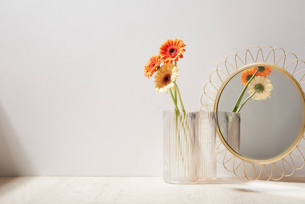 stylish mirror and glass vases with gerbera flowers on wooden surface in light room, close view - Photo, image