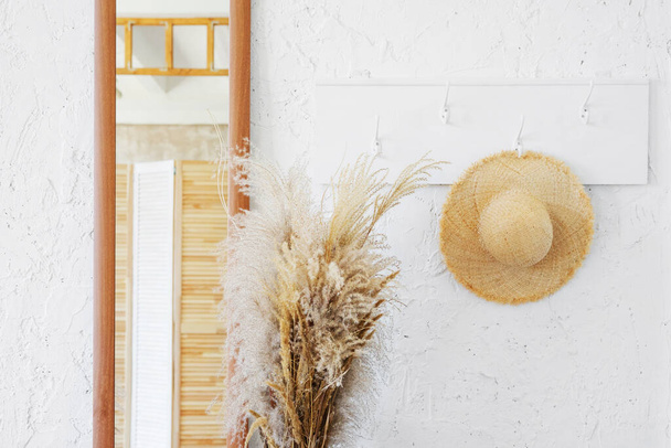 Wicker hat on a white wooden hanger in a minimalistic interior. A mirror and a bouquet of dry spikelets in the hallway of the Provencal cottage. - Foto, imagen