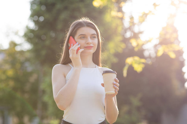 Ritratto di bella donna in piedi nel parco con smartphone e tazza di caffè tra le mani. - Foto, immagini