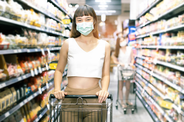 Asian woman wearing face mask push shopping cart in suppermarket . Girl choosing, looking somethings to buy at shelf during coronavirus crisis or covid19 .happy lifestyle or shopaholic concept. - Foto, imagen