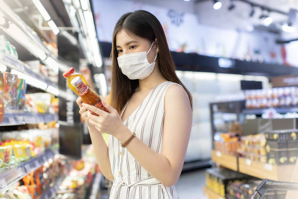 Asian woman wearing face mask push shopping cart in suppermarket . Girl choosing, looking somethings to buy at shelf during coronavirus crisis or covid19 .happy lifestyle or shopaholic concept. - Photo, image
