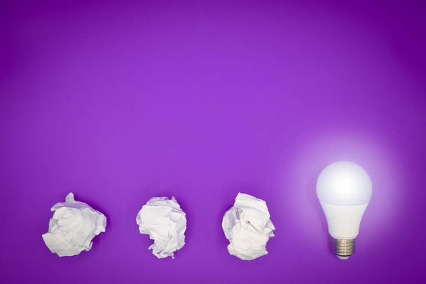 Crumpled paper balls and a light bulb glows. Copy space on purple background. Creativity, several attempts and successful idea concept. - Photo, Image