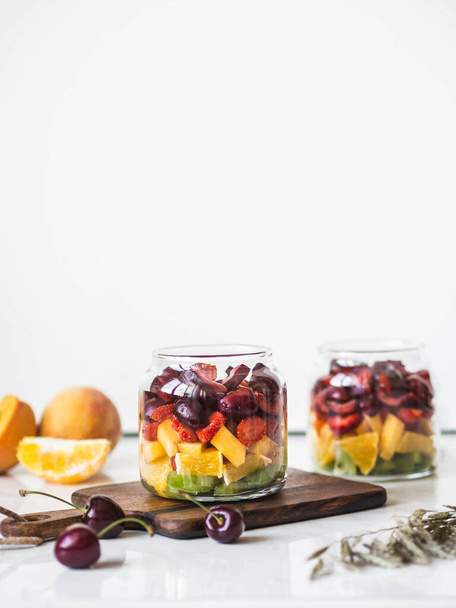 Two glass jars with berries and fruits. Fruit salad with kiwi, peach, strawberry, cherries and plum in glass jars on wood board. Summer seasonal tasty vegetarian salad for a snack or lunch - Fotografie, Obrázek