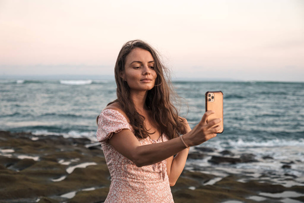 Красива дівчина перешкоджає дивлячись на телефон на пляжі на тлі моря. Гарна молода дівчина вирівнює волосся, дивлячись на мобільний телефон
. - Фото, зображення
