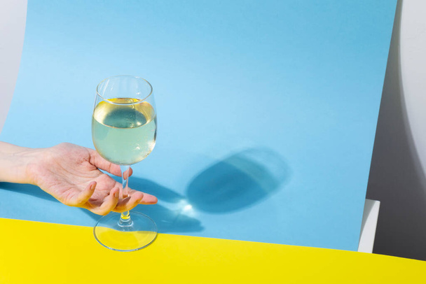 Female hand holds glass of white wine on yellow blue background, long shadow from the glass. Shadow effect, hard light - Foto, Imagem
